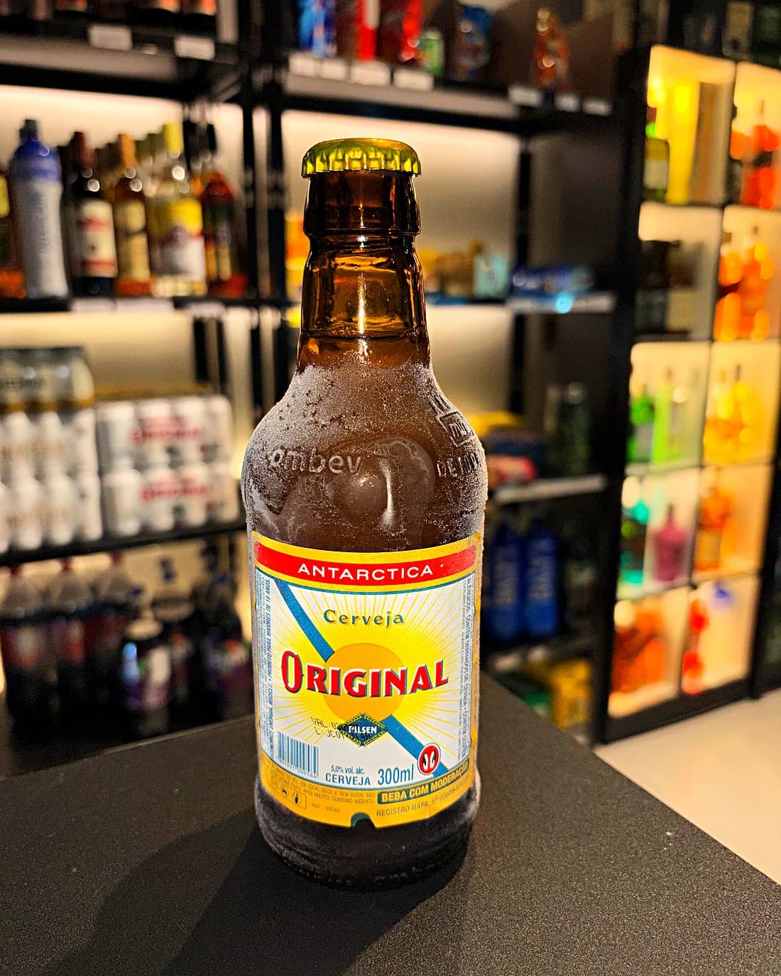 Garrafa gelada de Antarctica Original 300 ml no balcão, com as prateleiras ao fundo